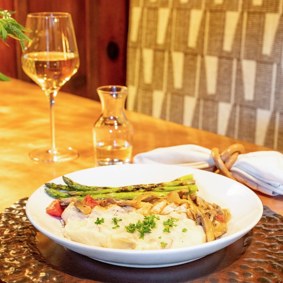 A white plate of mashed potatoes, chicken, mushrooms, asparagus, and tomatoes with wine.
