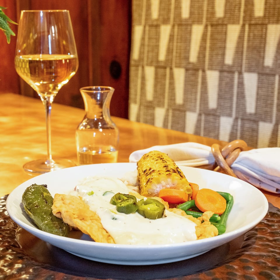 A white plate with a meal, including grilled corn, vegetables, and a creamy sauce, next to a glass of white wine.