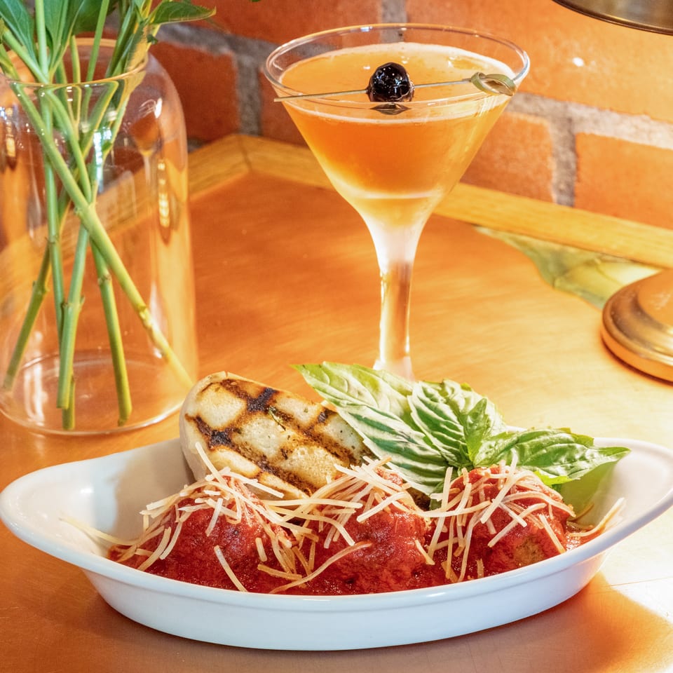 Meatballs in red sauce with parmesan, grilled bread, basil, and a cocktail on a wooden table.