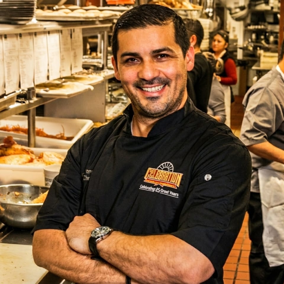 Smiling chef in a black Celebration jacket with crossed arms in a busy commercial kitchen.