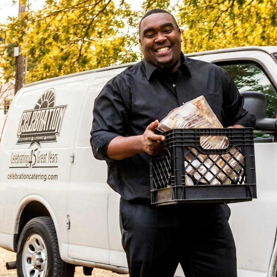Smiling Celebration Catering employee holds a crate of food next to a white catering van.